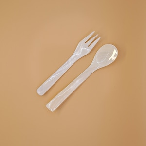 May include: A white plastic fork and spoon set. The fork has three tines, and the spoon has a rounded bowl. Both utensils have long, slender handles. The set is displayed on a solid beige background.