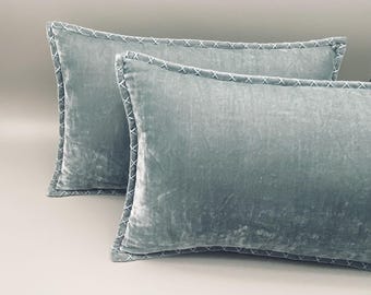 May include: Two rectangular throw pillows in a soft, dusty blue velvet fabric. Each pillow features a decorative border with a white cross-stitch pattern along the edges, providing a distinct contrast to the smooth, light-reflecting velvet surface.