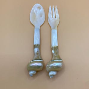 May include: A spoon and fork set. The spoon and fork heads are white with a pearlescent sheen. The handles are white, transitioning to a brown, shell-like base. The base has a spiral design and a small, pointed tip.
