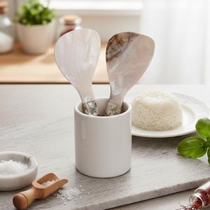 May include: Two white and iridescent rice paddles with decorative handles are in a white ceramic holder. A small scoop of salt and cooked rice are also visible, suggesting a kitchen or dining setting.