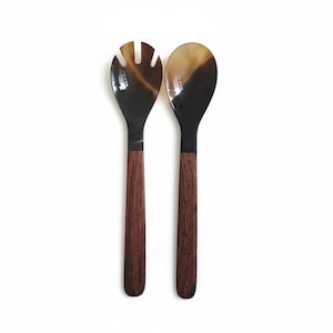 May include: A pair of salad servers. One is a spoon, the other a fork. The serving utensils have dark brown and tan tops with dark brown wooden handles. The servers are set against a white background.