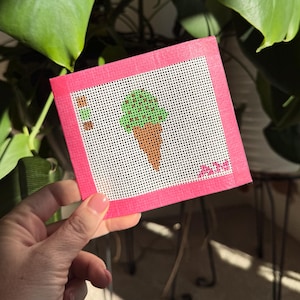 May include: A pink square needlepoint featuring a green ice cream cone with a brown cone. The needlepoint is held in front of a lush green plant. The needlepoint has a white background with a pink border.