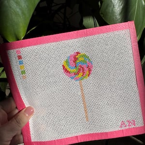 May include: A hand-stitched needlepoint canvas featuring a colorful lollipop design. The lollipop has a spiral pattern in shades of pink, yellow, blue, and green. The canvas is bordered with a bright pink edge and has a white background.