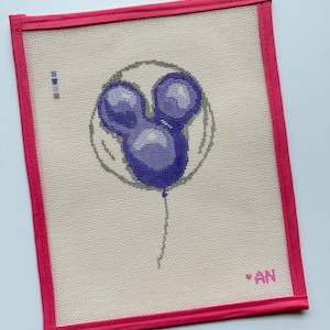 May include: Needlepoint canvas featuring a purple Mickey Mouse balloon design. The balloon is centered on a beige canvas, framed by a bright pink border. The initials "AN" are in the lower right corner.