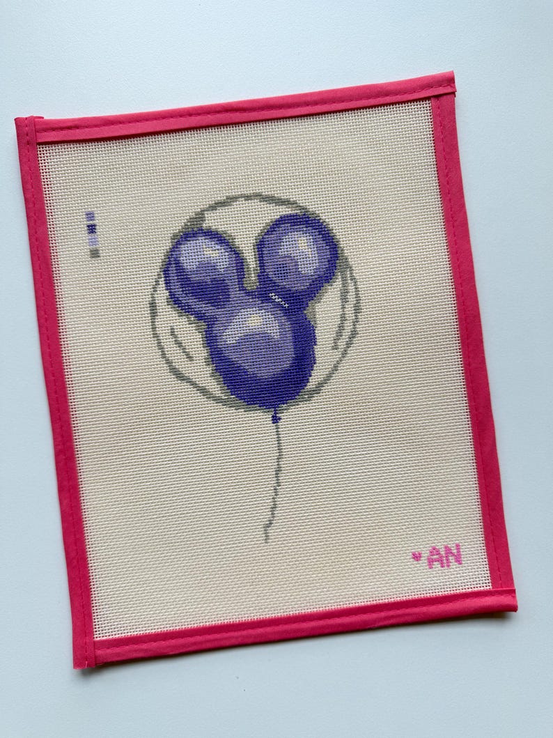 May include: Needlepoint canvas featuring a purple Mickey Mouse balloon design. The balloon is centered on a beige canvas, framed by a bright pink border. The initials "AN" are in the lower right corner.