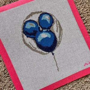 May include: Needlepoint canvas featuring a design of three blue balloons with highlights, set against a white background. The balloons are within a gray circle, and the canvas has a bright pink border. The canvas is on a brown, textured surface.