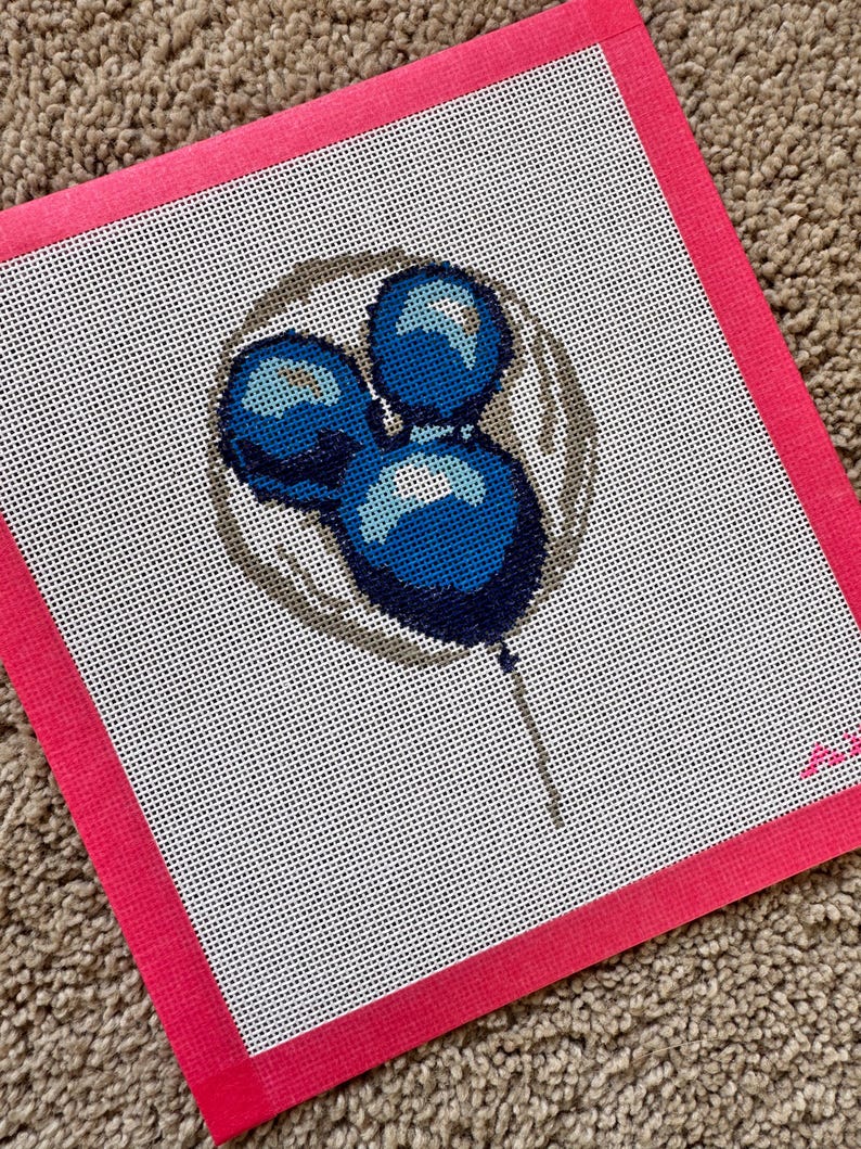 May include: Needlepoint canvas featuring a design of three blue balloons with highlights, set against a white background. The balloons are within a gray circle, and the canvas has a bright pink border. The canvas is on a brown, textured surface.