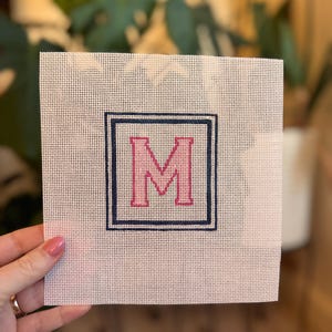May include: A square needlepoint canvas with a navy blue border and a pink letter "M" in the center. The canvas is held by a hand, showcasing the detailed stitching and design. The background is blurred, highlighting the canvas.