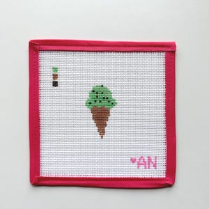 May include: A square needlepoint canvas featuring a cross-stitched mint green ice cream cone with brown chocolate chips. The canvas has a bright pink fabric border and includes a small color key on the left and the letters AN in the bottom right corner.