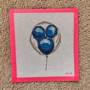 May include: A needlepoint canvas featuring a design of three blue balloons in a circle, with a string. The canvas is framed with a bright pink border. The background is white with a grid pattern. The design is stitched with various shades of blue.