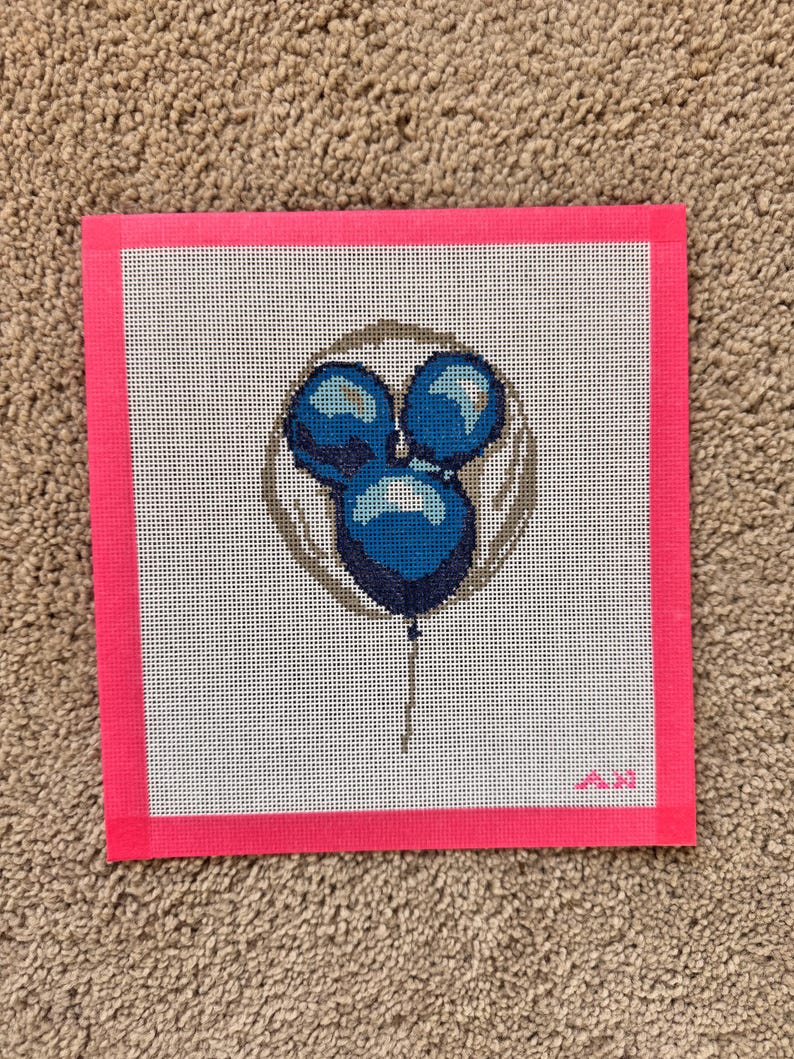 May include: A needlepoint canvas featuring a design of three blue balloons in a circle, with a string. The canvas is framed with a bright pink border. The background is white with a grid pattern. The design is stitched with various shades of blue.