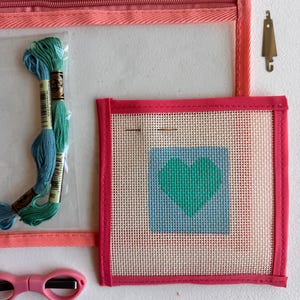 May include: A craft kit with a clear zippered pouch, featuring embroidery floss, a needlepoint canvas with a heart design, small pink scissors, and a metal tool. The canvas has a teal heart on a blue background.
