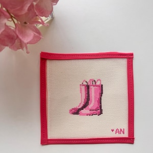 May include: A square, off-white needlepoint coaster with a pink border. The coaster features a cross-stitched design of pink rain boots. The initials "AN" are stitched in the lower right corner. Pink flowers are in the background.