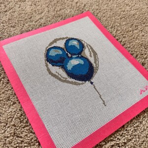 May include: A needlepoint canvas featuring a blue lollipop design. The lollipop is composed of three blue spheres with a brown outline, set against a white background with a pink border. The canvas is on a brown surface.