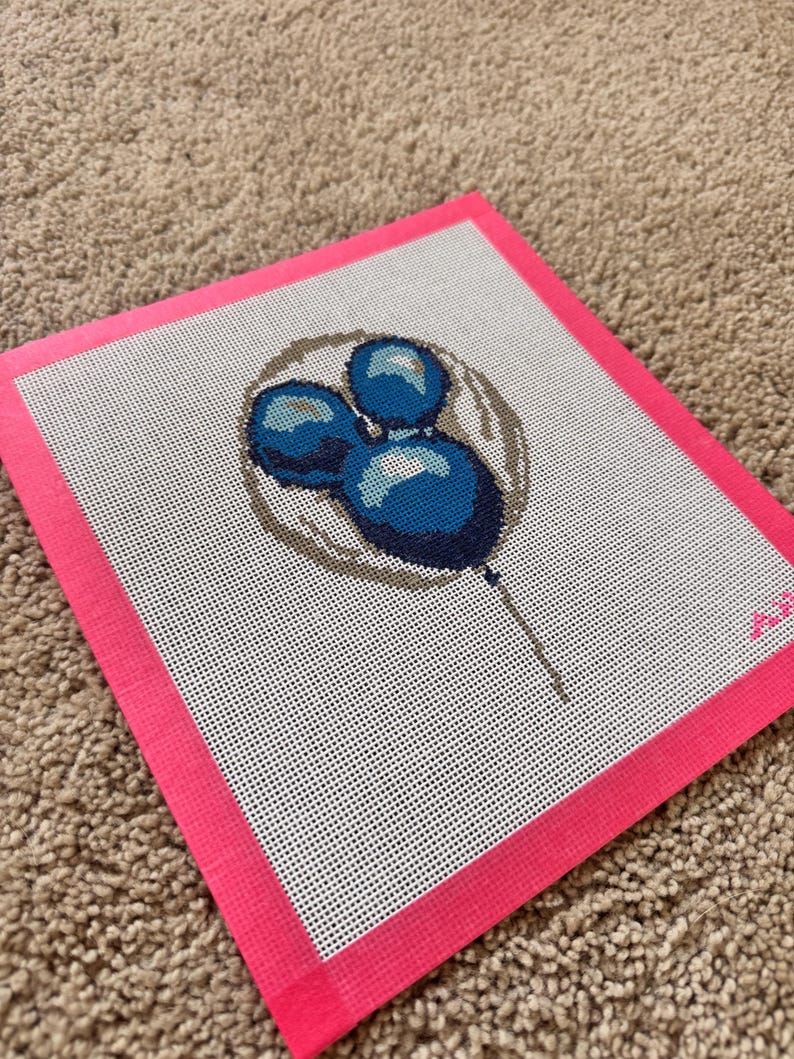 May include: A needlepoint canvas featuring a blue lollipop design. The lollipop is composed of three blue spheres with a brown outline, set against a white background with a pink border. The canvas is on a brown surface.