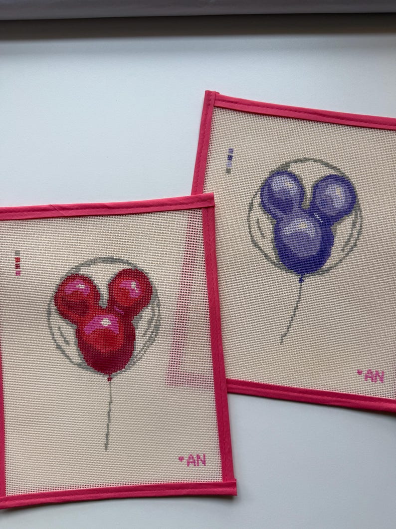 May include: Two needlepoint canvases with a Mickey Mouse balloon design. One balloon is red, the other is purple. Each canvas has a pink border and the initials "AN" in the corner.