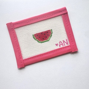 May include: A pink needlepoint coaster with a stitched watermelon slice design. The coaster has a white background and a pink border. The initials "AN" are stitched in pink on the right side of the coaster.