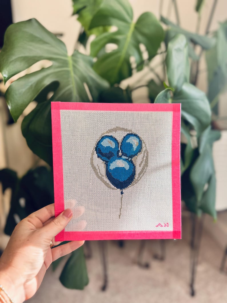May include: A needlepoint canvas featuring three blue balloons within a gray circle, with a pink border. The balloons are stitched in varying shades of blue, and the canvas is held in a hand. The background includes green foliage.