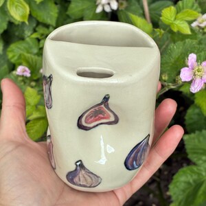 以下が含まれることがあります： クリーム色のセラミック花瓶、イチジクのデザイン。 花瓶は長方形で、上部が湾曲し、小さな穴があります。 イチジクは紫、赤、茶色の色合いで描かれています。