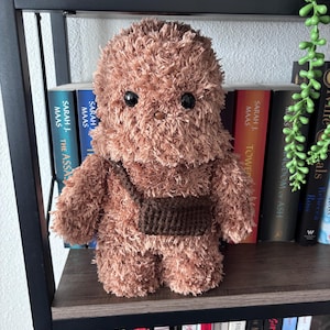 May include: A handmade, plush brown creature with black eyes and a small brown satchel. The stuffed animal is made of soft, textured material and is standing on a shelf in front of a collection of books.