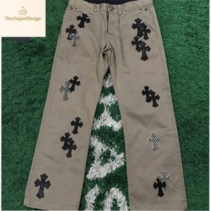 Może przedstawiać: Spodnie w kolorze khaki z wieloma czarnymi aplikacjami w kształcie krzyży. Niektóre krzyże mają wzór w szachownicę. Spodnie mają klasyczny krój z pięcioma kieszeniami oraz zapięcie na guzik i zamek błyskawiczny. Widoczna jest nazwa marki "NewSuperDesign".