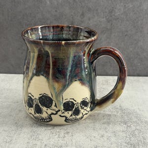 Puede incluir: Una taza de cerámica con base de color crema y dibujos de líneas negras de cráneos humanos en la parte inferior. La parte superior y el asa tienen un acabado con un esmalte brillante de varios tonos en colores marrón, azul y verde.