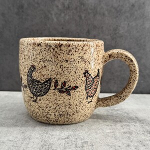 May include: A ceramic mug with a speckled beige finish featuring black line art illustrations of two chickens and a small leafy branch with red accents. The mug has a rounded body and a curved handle, showcasing a textured, earthy appearance.