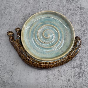 Könnte beinhalten: Eine Keramikschale in Schneckenform mit einem braunen, gesprenkelten Körper und einem kreisförmigen, hellblauen Gehäuse mit einem erhabenen Spiralmuster in der Mitte. Die Schale hat zwei kleine, abgerundete Fühler am Kopf der Schneckenbasis.