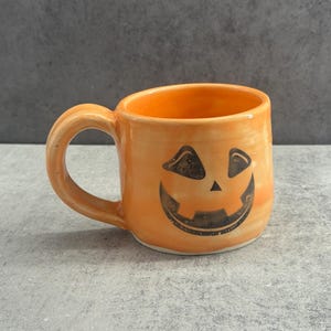 Peut inclure: Un mug en céramique orange brillant orné d'un visage de citrouille d'Halloween peint en noir, avec des yeux triangulaires, un petit nez triangulaire et un large sourire édenté. Le mug présente un corps arrondi et une anse incurvée.