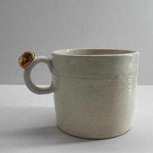 May include: Cream-coloured ceramic mug with a gold-coloured accent on the handle. The mug has a cylindrical shape and a simple, elegant design. The handle is a loop with a decorative gold embellishment.