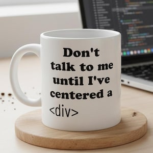 May include: White ceramic mug with the text "Don't talk to me until I've centered a <div>" in black. The mug sits on a wooden coaster. A laptop with code on the screen is in the background.