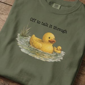 May include: Olive green t-shirt featuring a yellow rubber duck and duckling in water, with the text "Off to talk it through." The graphic is a whimsical illustration. The shirt is folded on a wooden surface.
