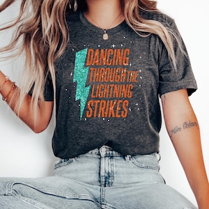 Puede incluir: Camiseta gris oscuro con el texto "DANCING THROUGH THE LIGHTNING STRIKES" en naranja y un rayo de purpurina turquesa. La camiseta es de cuello redondo y tiene mangas cortas.