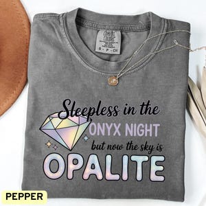 Pode incluir: Uma camiseta cinza Comfort Colors com o texto "Sleepless in the Onyx Night but now the sky is Opalite" e um gráfico de diamante. Um colar de prata com um pingente está sobre a camiseta. A camiseta está rotulada como "Pepper".