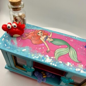 May include: A decorative, rectangular box with a pink top featuring a mermaid illustration. The box is painted light blue with glitter accents. A small red crab figurine and a glass bottle filled with seashells sit on top. The box has two small windows.