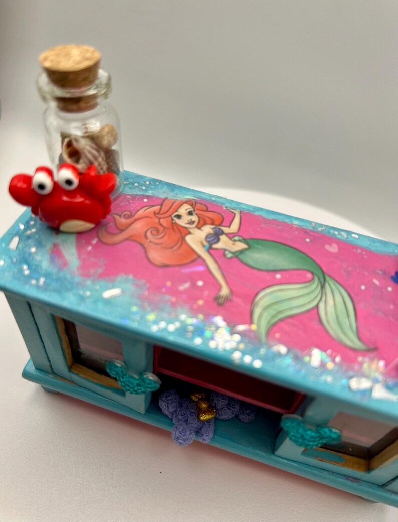 May include: A decorative, rectangular box with a pink top featuring a mermaid illustration. The box is painted light blue with glitter accents. A small red crab figurine and a glass bottle filled with seashells sit on top. The box has two small windows.