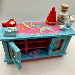May include: A miniature, turquoise cabinet with a pink and blue top featuring a mermaid illustration. The cabinet has opening doors with Mickey Mouse-shaped handles. Accessories include a red crab, a book titled "Moby Dick", and a glass jar.
