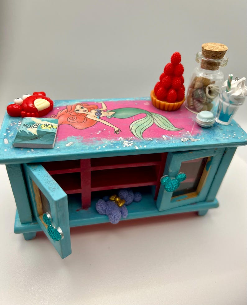 May include: A miniature, turquoise cabinet with a pink and blue top featuring a mermaid illustration. The cabinet has opening doors with Mickey Mouse-shaped handles. Accessories include a red crab, a book titled "Moby Dick", and a glass jar.