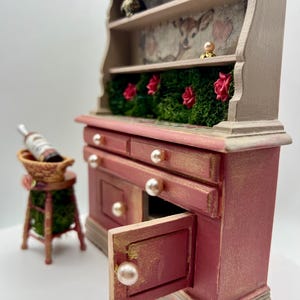 May include: A miniature, hand-painted, red and gold hutch with pearl knobs and a small stool. The hutch has shelves with a deer illustration, red roses, and a small frog figurine. A bottle of wine sits in a basket on the stool.