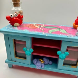 May include: A small, light blue wooden cabinet with a painted image of Ariel from The Little Mermaid. The cabinet has two doors with Mickey Mouse-shaped embellishments and a red interior. A red crab figurine and a glass bottle with a cork stopper are on top.