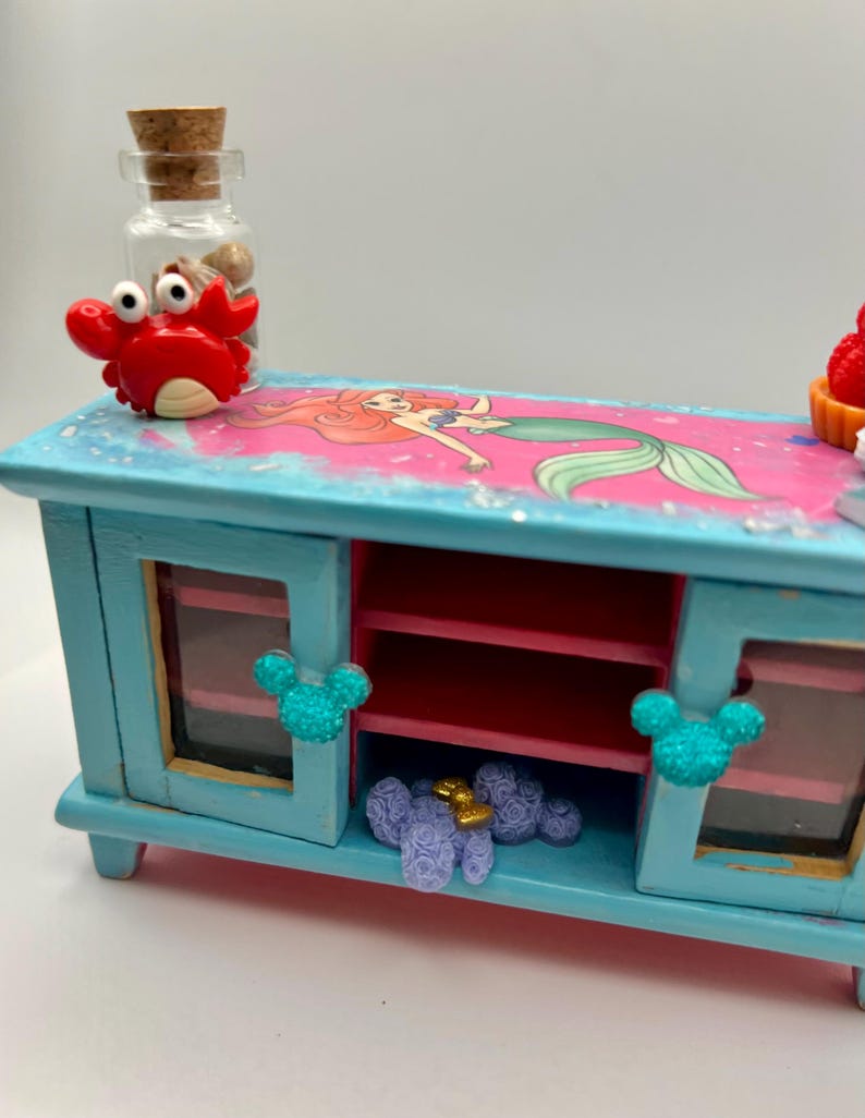 May include: A small, light blue wooden cabinet with a painted image of Ariel from The Little Mermaid. The cabinet has two doors with Mickey Mouse-shaped embellishments and a red interior. A red crab figurine and a glass bottle with a cork stopper are on top.