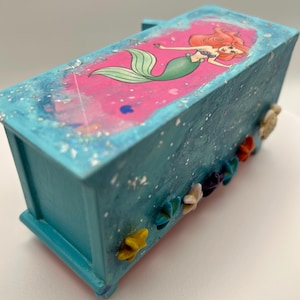 May include: A turquoise decorative box featuring a pink top with a mermaid illustration. The box is adorned with colorful flower and seashell embellishments. The mermaid has red hair and a green tail.