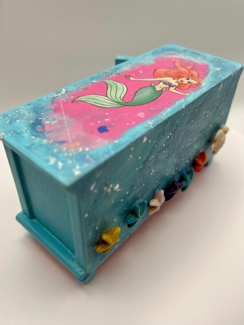 May include: A turquoise decorative box featuring a pink top with a mermaid illustration. The box is adorned with colorful flower and seashell embellishments. The mermaid has red hair and a green tail.