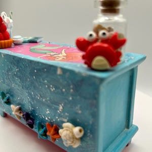 May include: A small, light blue decorative chest with a mermaid illustration on top. The chest is adorned with starfish and turtle embellishments. A red crab figurine and a small glass bottle with a cork stopper are on top.