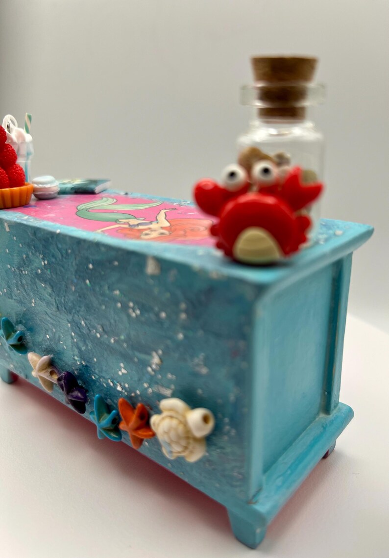 May include: A small, light blue decorative chest with a mermaid illustration on top. The chest is adorned with starfish and turtle embellishments. A red crab figurine and a small glass bottle with a cork stopper are on top.