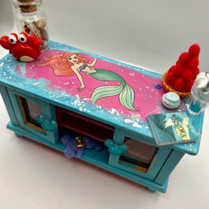 May include: A miniature, hand-painted blue cabinet with a pink and blue top featuring an Ariel the Little Mermaid graphic. The cabinet is decorated with a red crab, a bottle of shells, and a miniature dessert. The cabinet has two windows and a shelf.