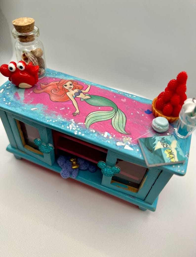 May include: A miniature, hand-painted blue cabinet with a pink and blue top featuring an Ariel the Little Mermaid graphic. The cabinet is decorated with a red crab, a bottle of shells, and a miniature dessert. The cabinet has two windows and a shelf.