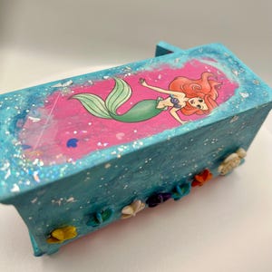 May include: A small, rectangular, turquoise box with a pink interior featuring a cartoon mermaid illustration. The box is decorated with glitter and small, colorful seashell embellishments, creating a whimsical, decorative item.