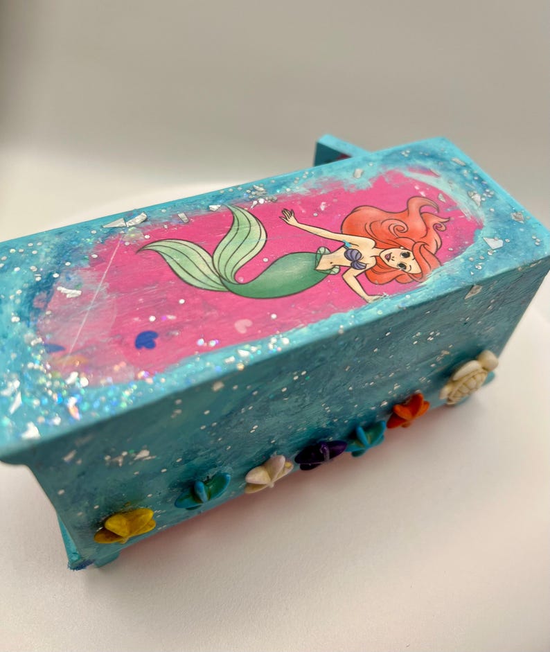 May include: A small, rectangular, turquoise box with a pink interior featuring a cartoon mermaid illustration. The box is decorated with glitter and small, colorful seashell embellishments, creating a whimsical, decorative item.