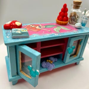 May include: A miniature, hand-painted, turquoise cabinet with a pink and blue top featuring a mermaid illustration. The cabinet has two doors with Mickey Mouse-shaped handles, and is decorated with miniature food items.